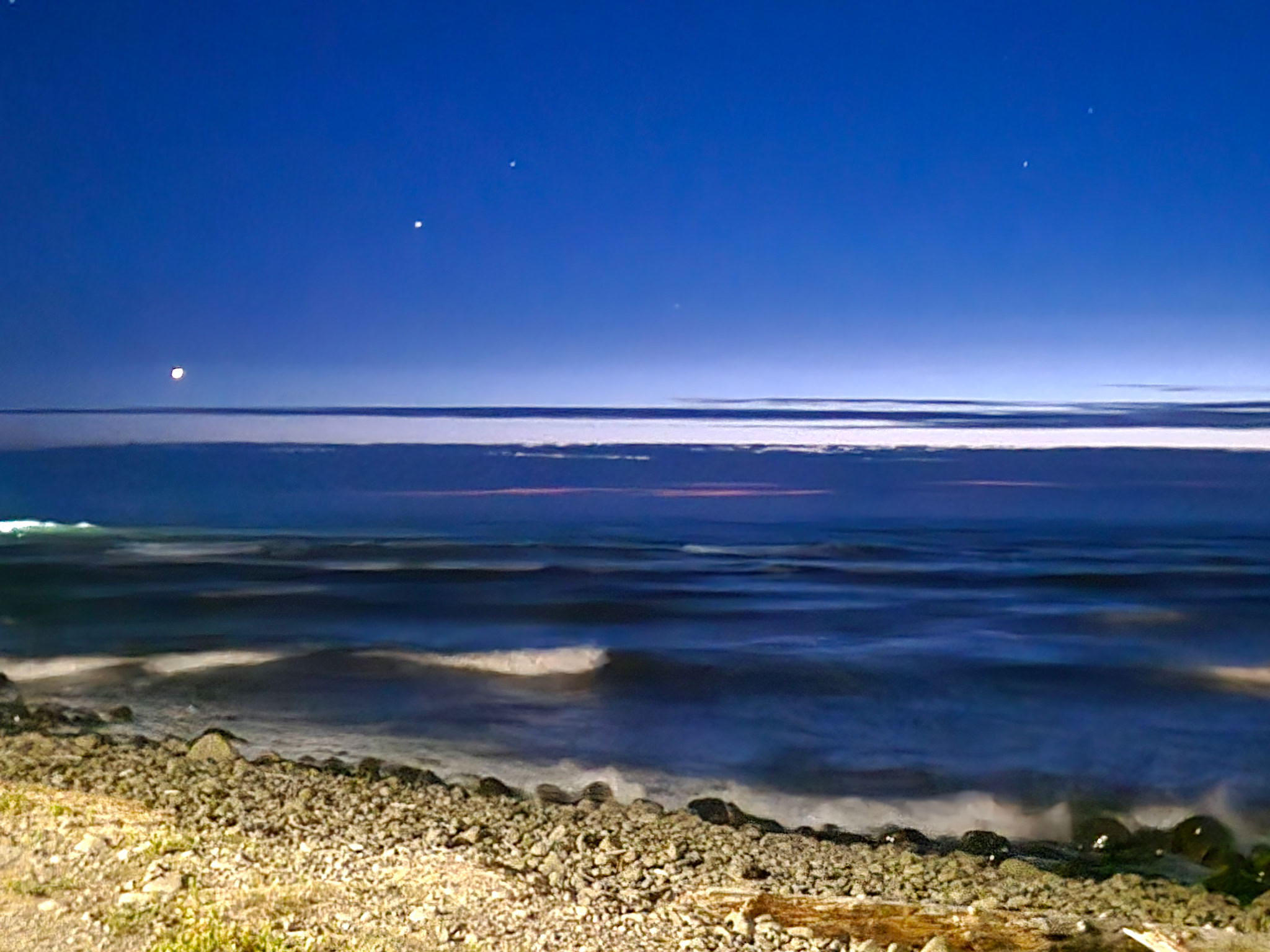 night time view of the ocean, sitting in the car with the lights illuminating the rocky ground between the viewer and the waves. the sky is deep and the stars are beginning to come out. the whole video is blurred and dark like a memory