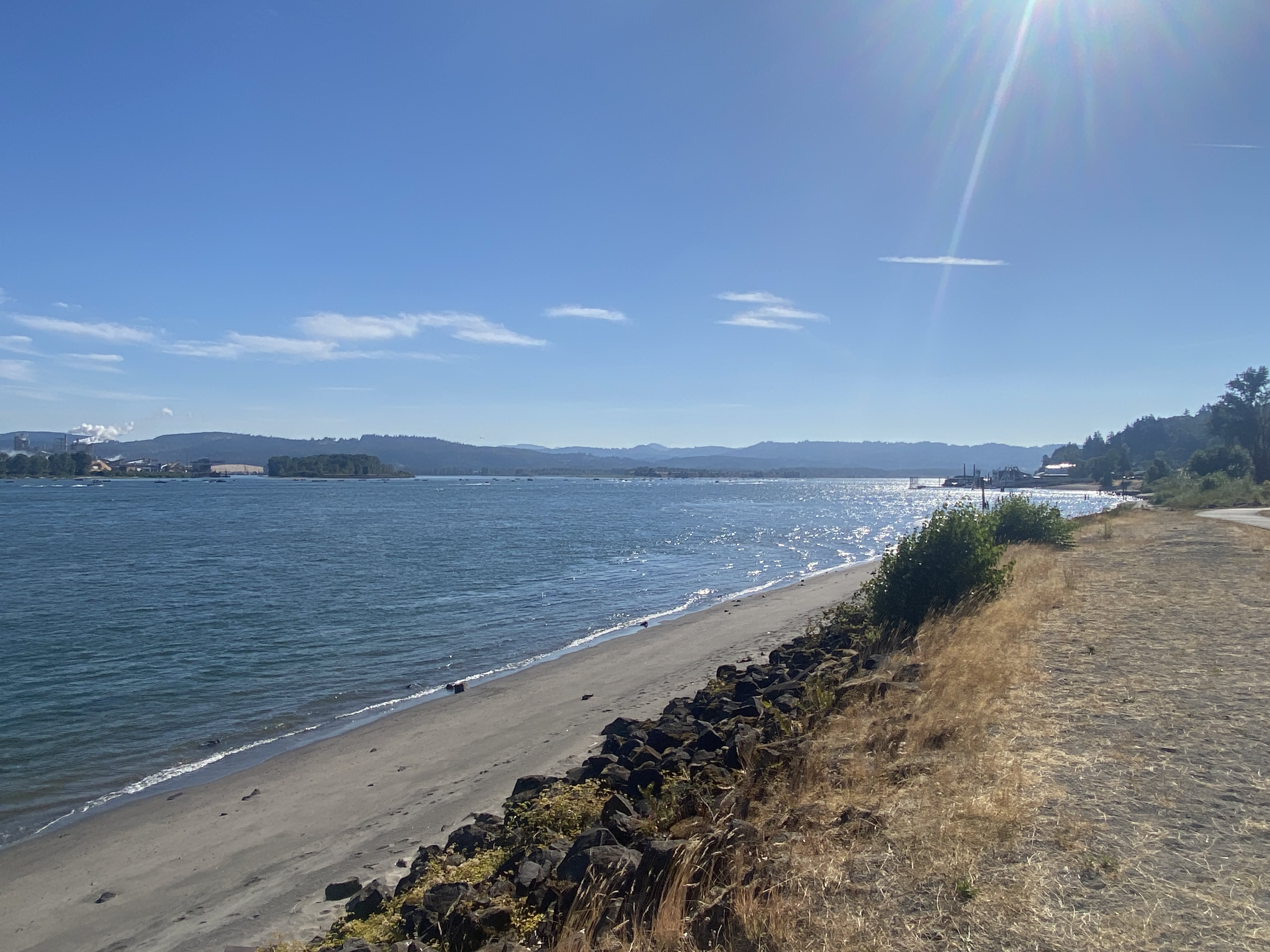 viewer is standing above the shore of a massive river. the sun is streaming into the photo, while hundreds of small boats dot the giant river. Across the river is foothills filled with trees, and a large port
